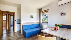 A modern room with a wooden floor, a blue sofa, a dining table with chairs, a window with curtains, and a small kitchen area. - Apartment "Gartenheim Martin" with Mountain View, Pool & Sauna (Scena)
