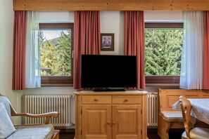 Une pièce avec un téléviseur posé sur un meuble en bois, une chaise, une table et une vue sur les arbres à travers de grandes fenêtres.