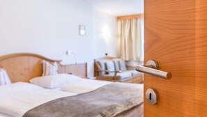 A hotel room with a wooden bed, a bedside table, a lamp, and a window with curtains. - Apartment 'Prapoz Typ B1' with Mountain View, Shared Garden & Wi-Fi (Ortisei)