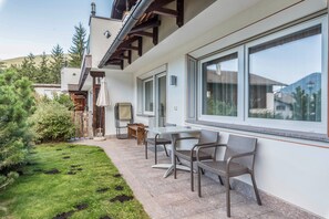 Une terrasse avec une table et des chaises, un banc en bois et une grande fenêtre donnant sur un jardin et une montagne.