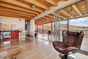 A spacious interior with a wooden ceiling, a dining area with chairs, a bar area with a counter, and a large window offering a view of the desert landscape.