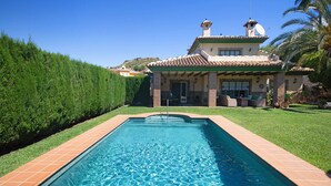 Uma piscina retangular com borda azulejada, um espaço de pátio com mobiliário de exterior e uma casa com telhado e antena parabólica.