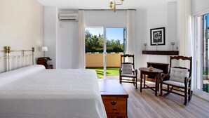 Quarto de hotel com uma cama, um baú de madeira, uma cadeira, uma mesa pequena, lareira e vista para o exterior.