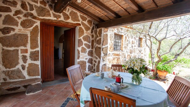 A stone-walled room with a wooden ceiling, a round table with chairs, and a view of a garden. - Holiday Home "Sa Domedda" with Mountain View, Garden & A/C (Orosei)