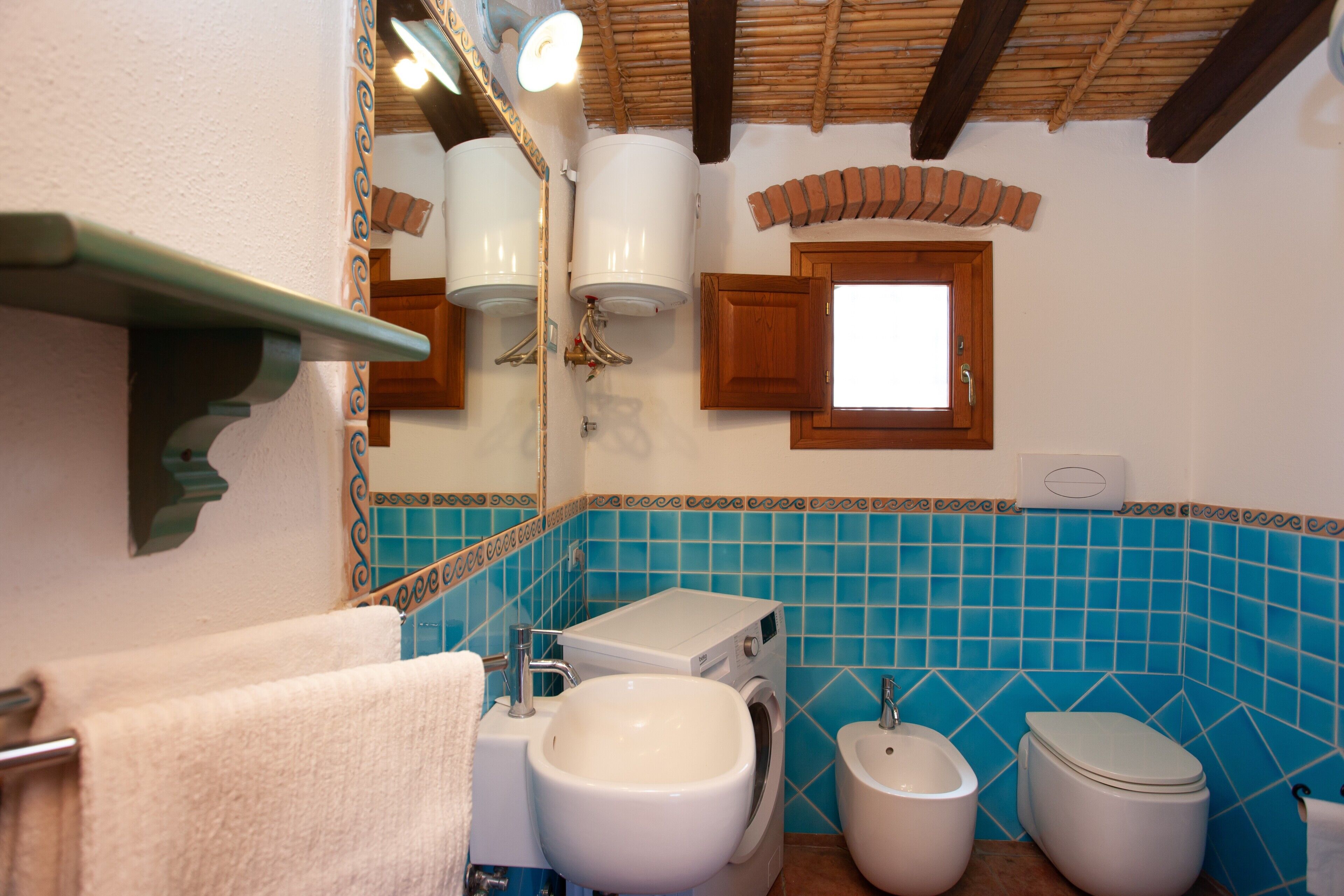 A bathroom with a sink, toilet, and washing machine, a mirror, and a shelf with a towel.