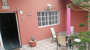 Een kamer met tegels op de vloer, een raam met tralies, een tafel met stoelen, een cactus in een pot, en een deur.