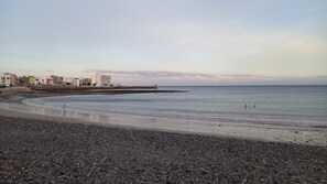 Uma praia de seixos com mar calmo, edifícios ao fundo e céu claro.