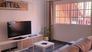 Een woonkamer met een flatscreen televisie, een bank, een bijzettafeltje en een raam met verticale jaloezieën.