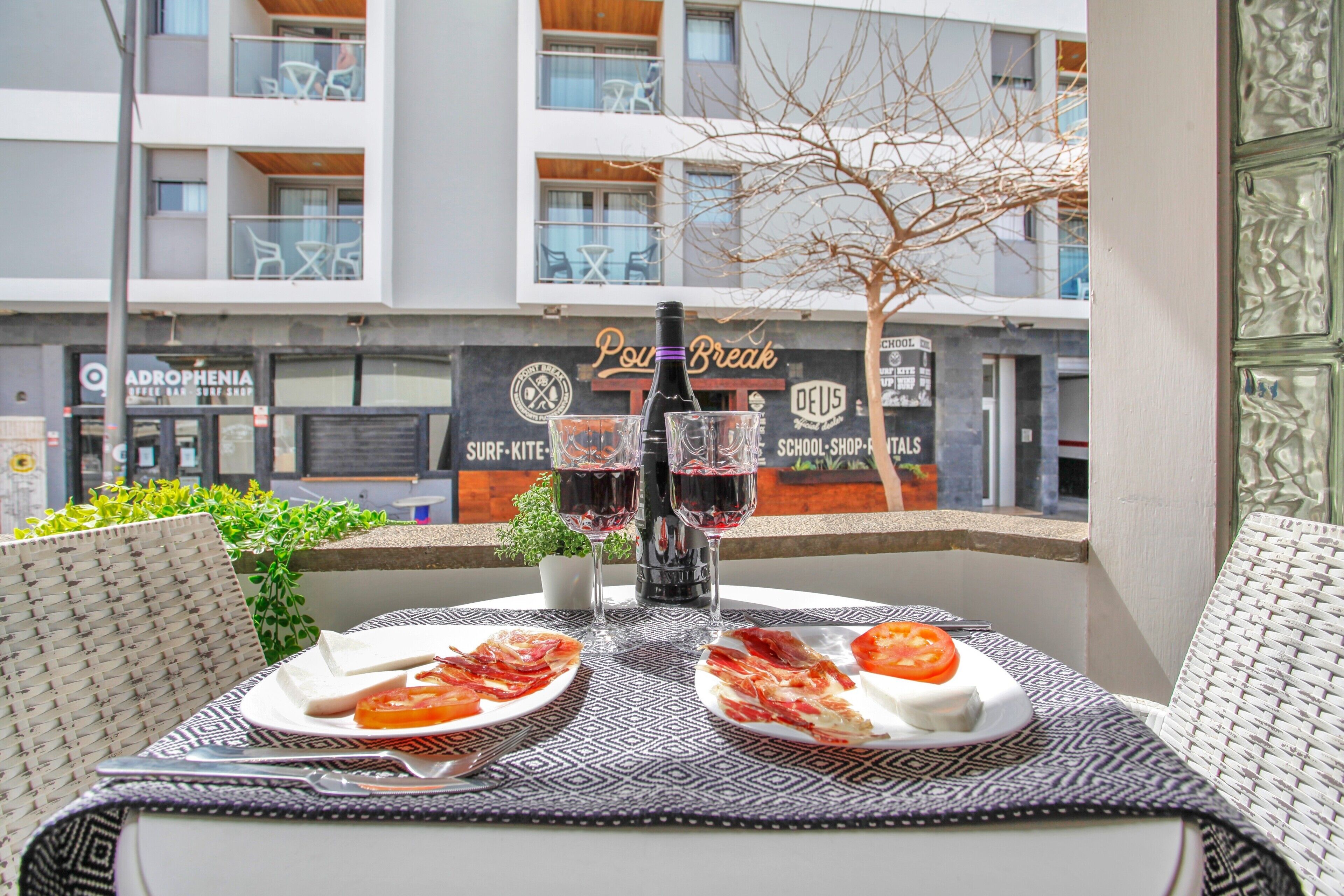 Ein Tisch mit Speisen, Weingläsern, einer Weinflasche und einem Menüständer auf einem Balkon mit Blick auf eine Stadtlandschaft und verschiedene Geschäfte.