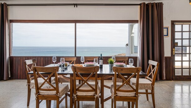 Un comedor con una mesa dispuesta para seis personas, sillas de madera, vista al mar y una puerta con panel de vidrio.