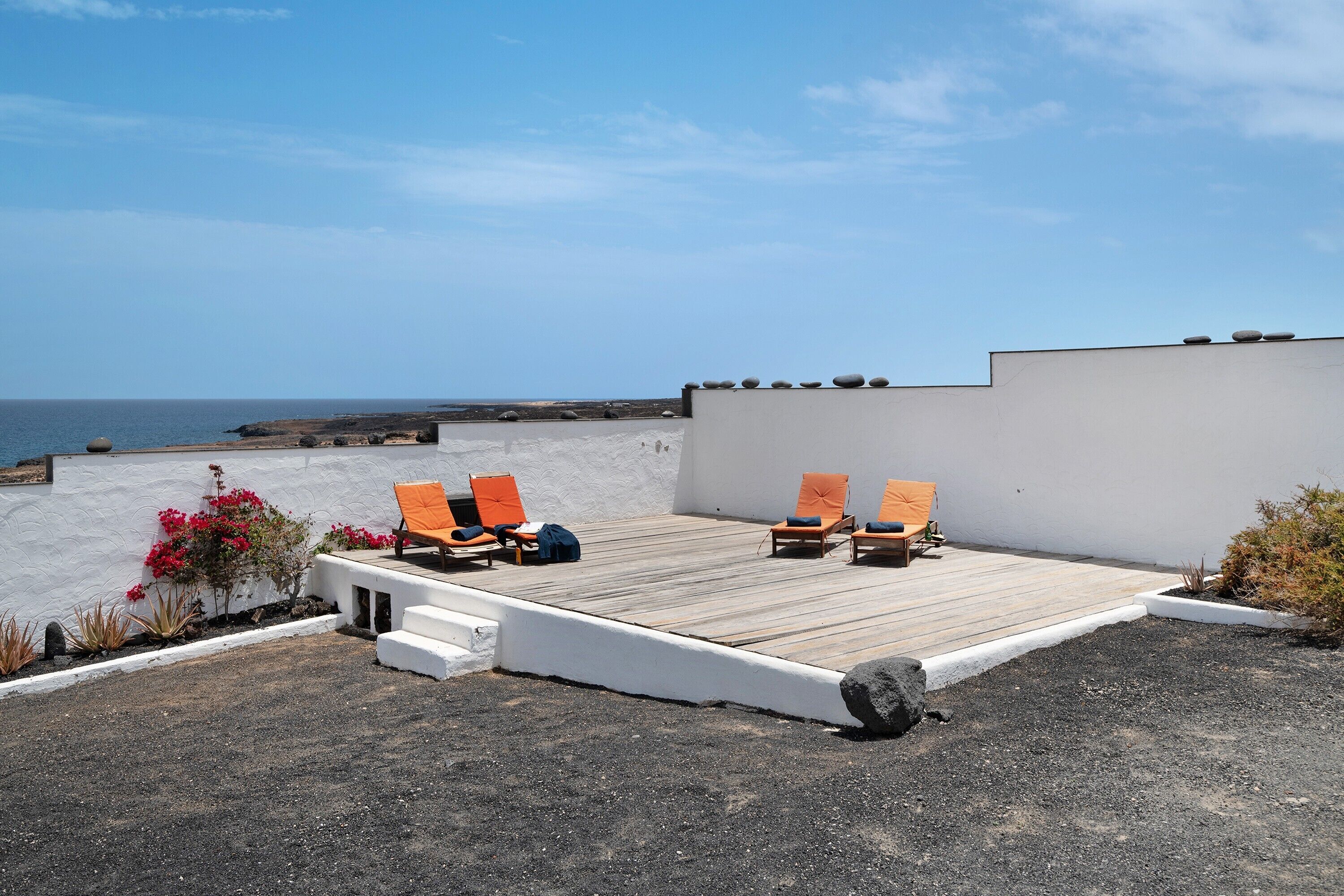 Terraza en la azotea con vistas al mar, que incluye una cubierta de madera, sillas naranjas y una pared blanca.