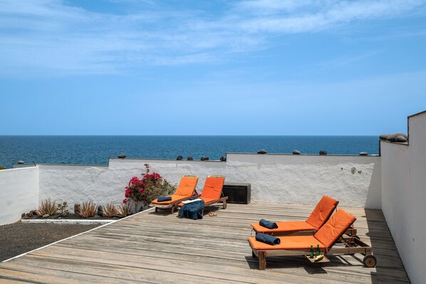 A rooftop terrace with wooden flooring, orange lounge chairs, and a view of the ocean.