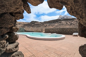 A swimming pool with a stone wall in the foreground, a white building on a hill in the background, and a clear sky.