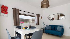 A modern living room with a dining table, chairs, a blue sofa, a large window with curtains, a hanging light fixture, and a wall-mounted mirror.