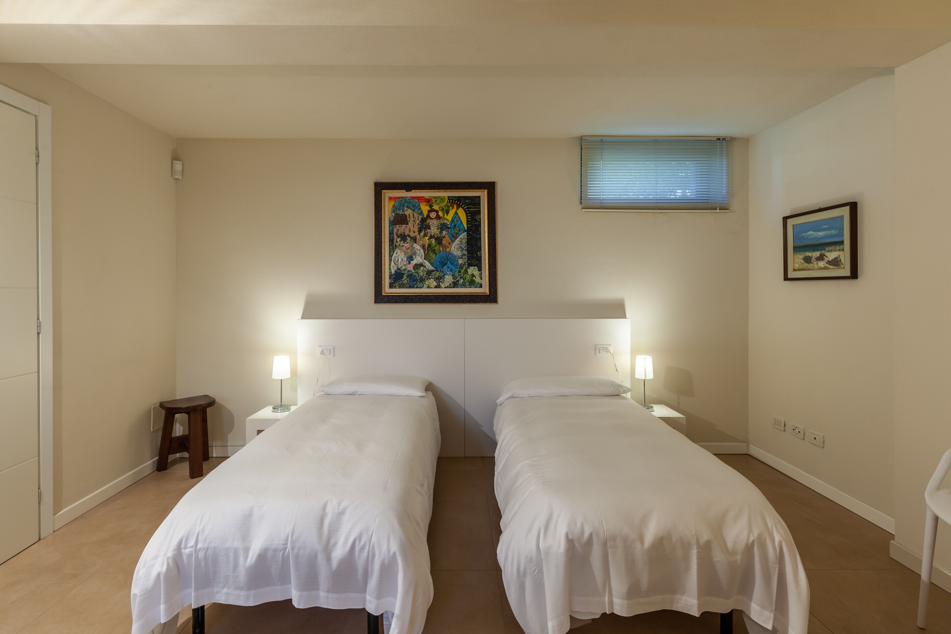 Ein Hotelzimmer mit zwei Einzelbetten, einem Nachttisch mit Lampe, einem kleinen Hocker und einem Bild an der Wand.