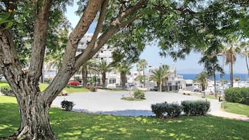 Une cour bordée d’arbres, d’où l’on aperçoit la mer et des bâtiments en arrière-plan.
