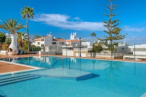 A swimming pool with clear blue water, surrounded by palm trees and a white building with red roofs.