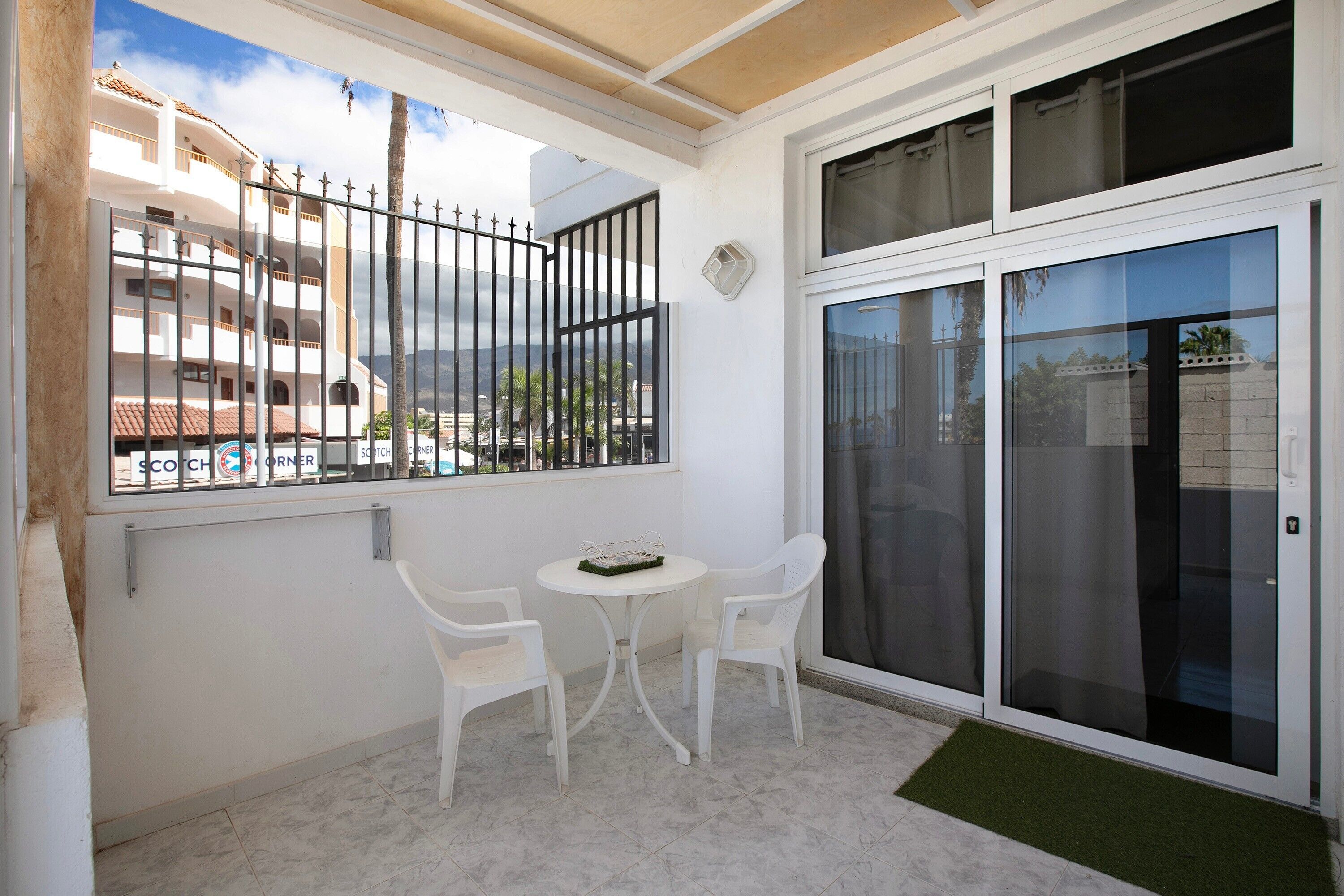 Un balcón con una mesa y sillas blancas, una puerta corrediza de cristal y vistas a un edificio con balcones.