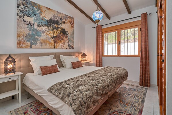 A bedroom with a large bed, a nightstand, a wall-mounted lamp, a window with curtains, and a painting.