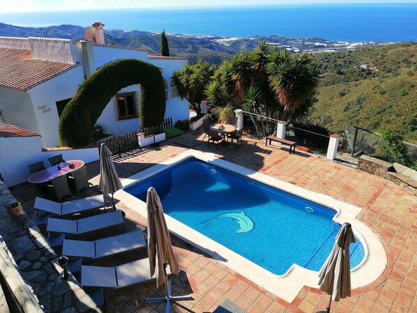 A swimming pool with a view of the sea, surrounded by a patio with chairs and umbrellas.