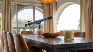 Uma sala de jantar com mesa, cadeiras, um telescópio e uma grande janela com cortinas.