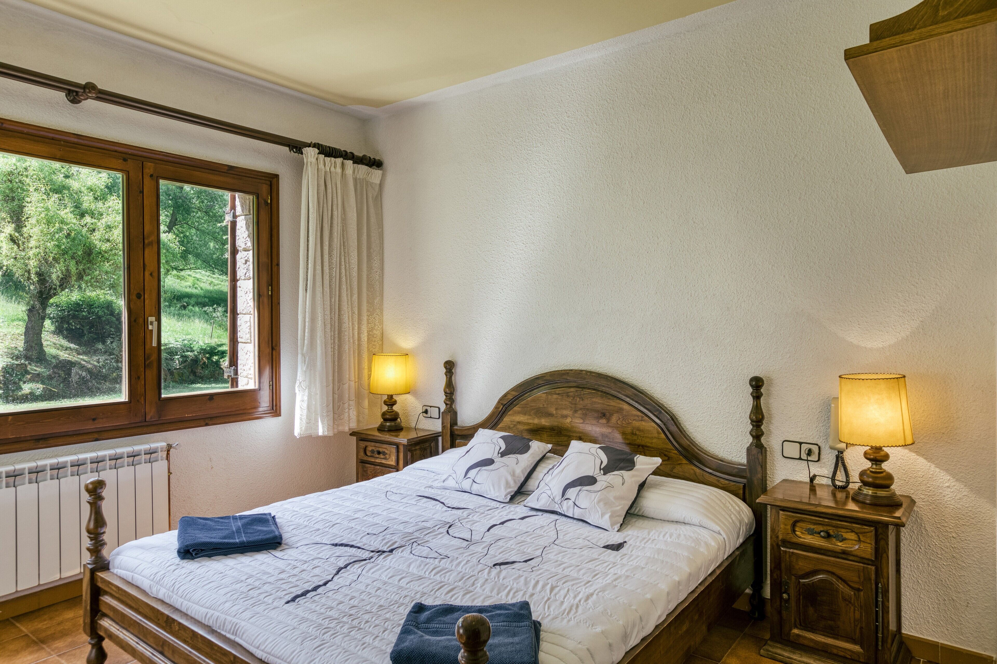 Un dormitorio con una cama de madera, dos lámparas de noche, un ventanal con cortinas y un radiador.