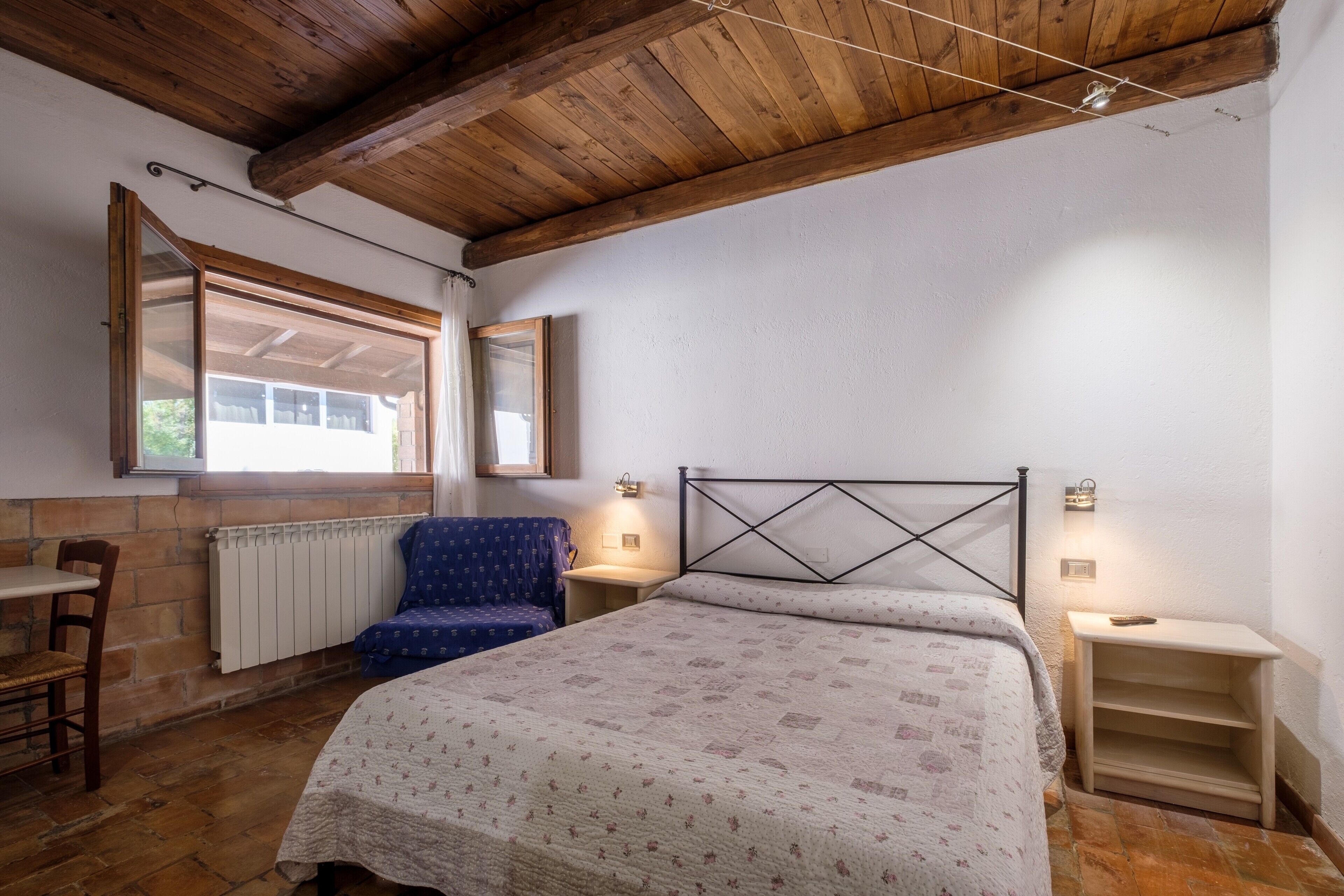 A bedroom with a wooden ceiling, a bed with a patterned bedspread, a chair, a nightstand, and a window with a view.
