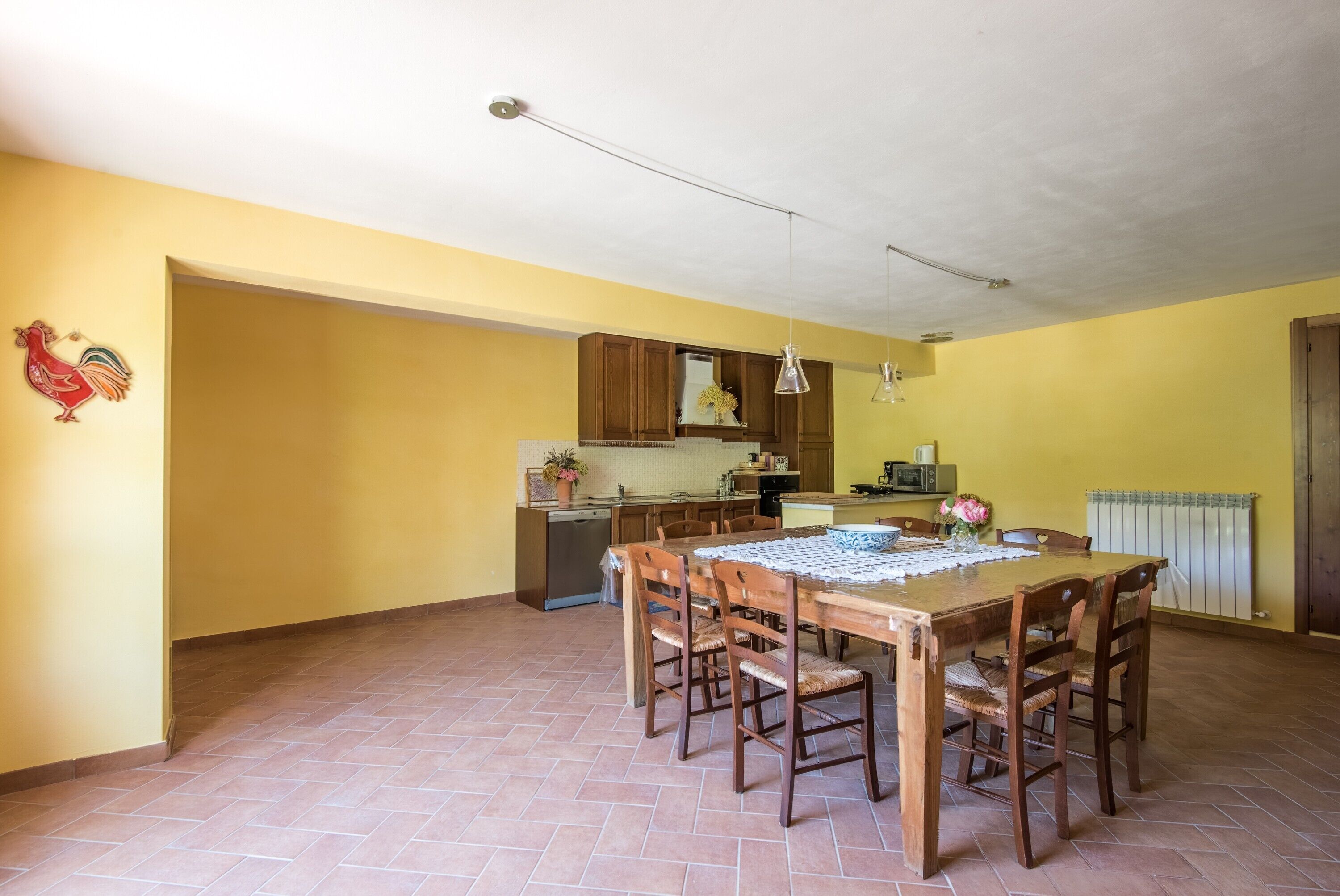 A dining area with a wooden table and chairs, a kitchen in the background, and a yellow wall.