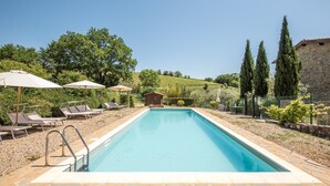 Una piscina rettangolare con bordi in pietra, circondata da lettini e ombrelloni, con un edificio in pietra e alberi sullo sfondo.