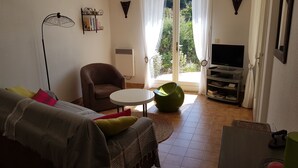 Een woonkamer met een bank, fauteuil, groene relaxstoel, tv-meubel en een raam met uitzicht op groen.