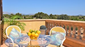Un balcon avec des fauteuils en osier et une table en verre, un saladier rempli de citrons et une vue sur des montagnes et des arbres lointains.