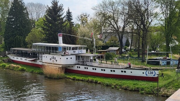 A riverboat with the number 120 on its side, docked at a pier with a grassy bank and trees in the background.