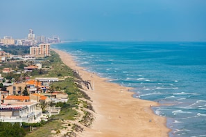 Paisagem costeira com uma praia de areia, edifícios e céu azul claro.
