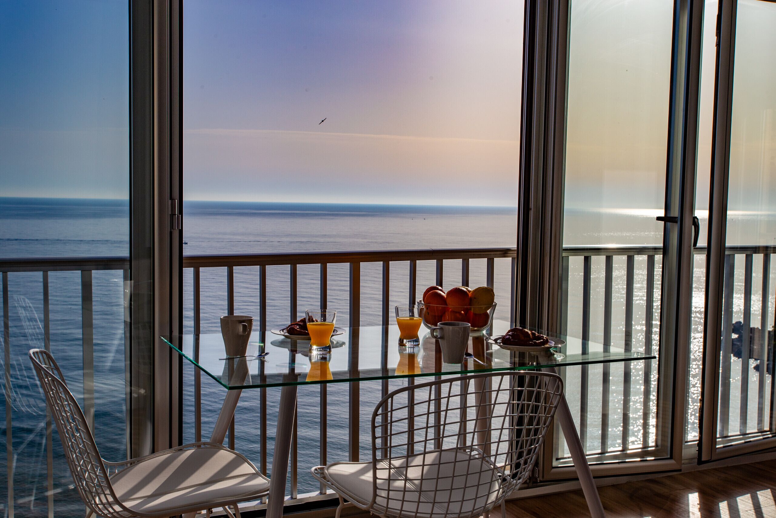 A balcony with a glass table, two chairs, and a view of the sea.