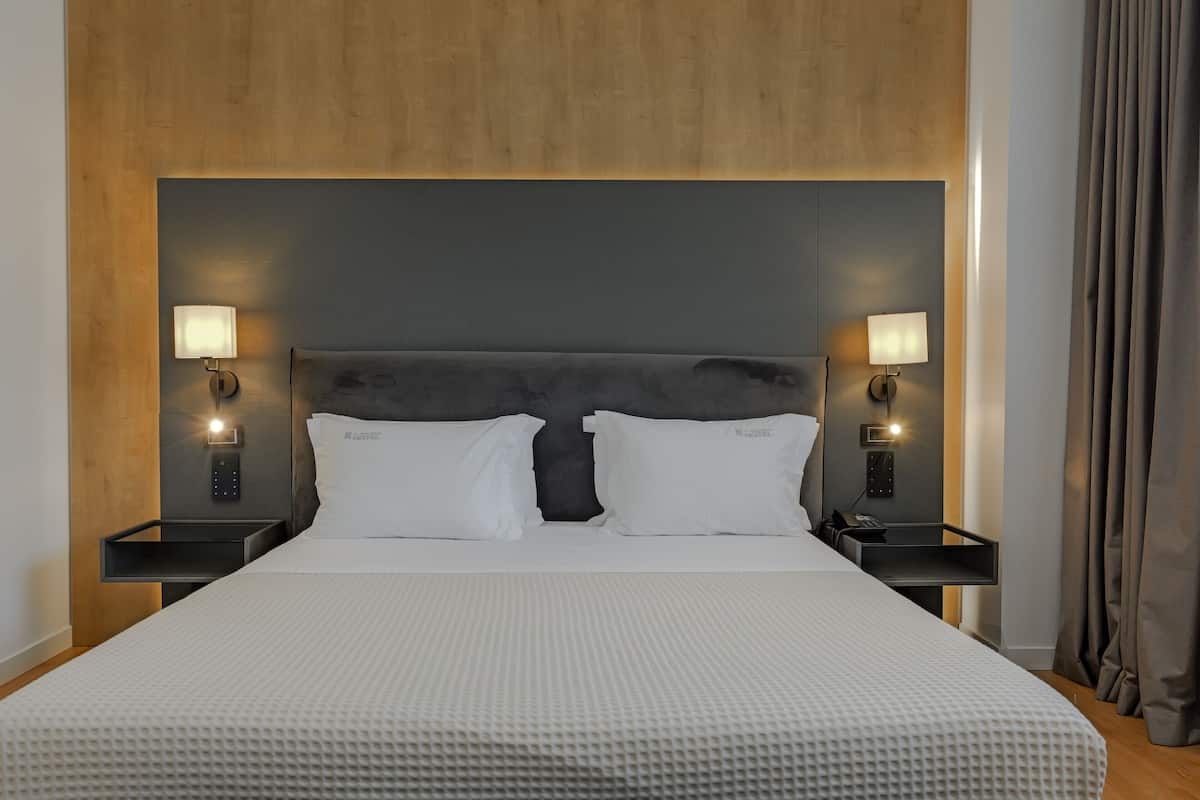 Moderni hotellihuone, jossa on suuri sänky, kaksi yöpöytää valaisimineen ja puinen sängynpääty.