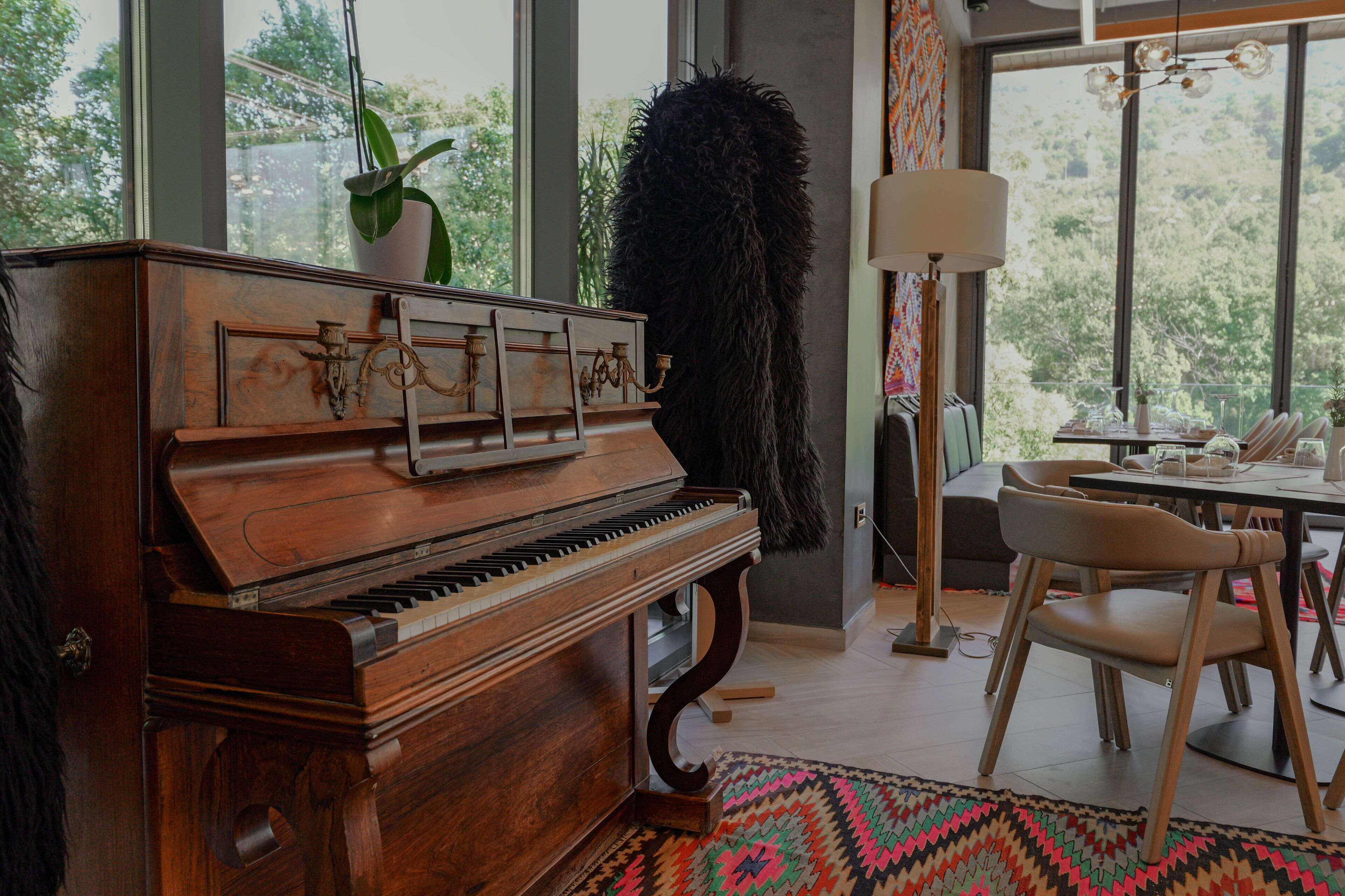 A grand piano in a well-lit room with large windows, a dining table, chairs, and a lamp.