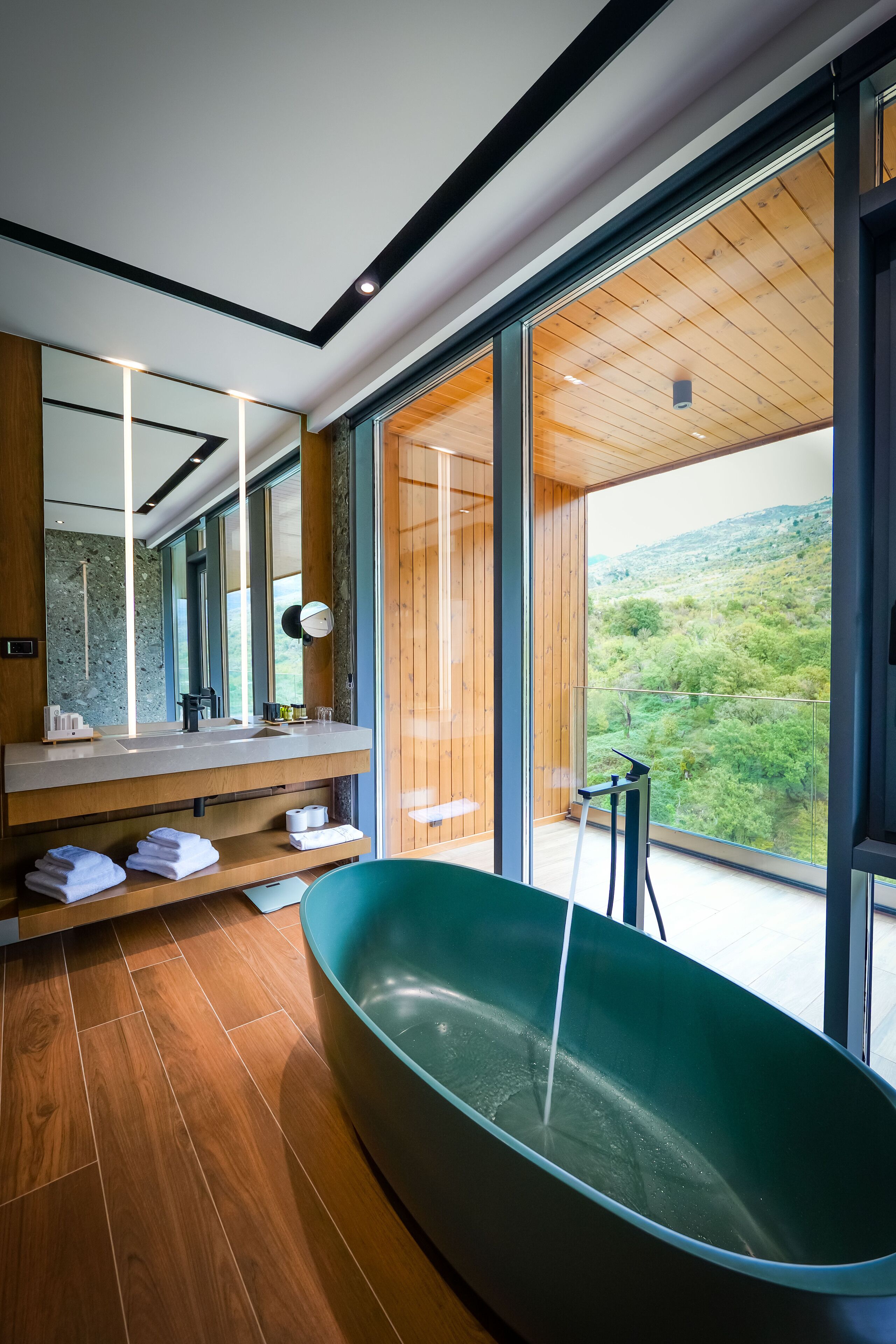 Un baño moderno con una bañera grande, suelo de madera, un espejo amplio y vistas a un paisaje verde.