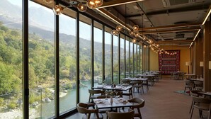 A restaurant with large windows offering a view of a river and mountains. Tables are set with chairs and centerpieces. - Uji i Ftohte Hotel (Qendër Tepelenë)