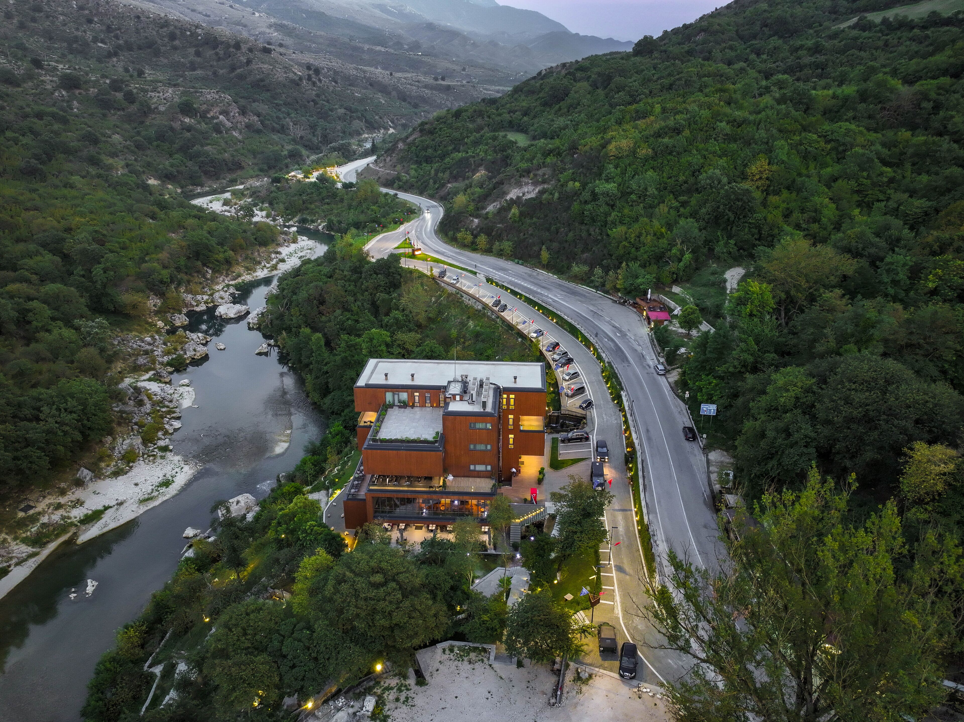 An aerial view of a modern hotel with a curved road leading to it, surrounded by lush greenery and a river.