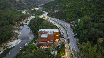 An aerial view of a modern hotel with a curved road leading to it, surrounded by lush greenery and a river.