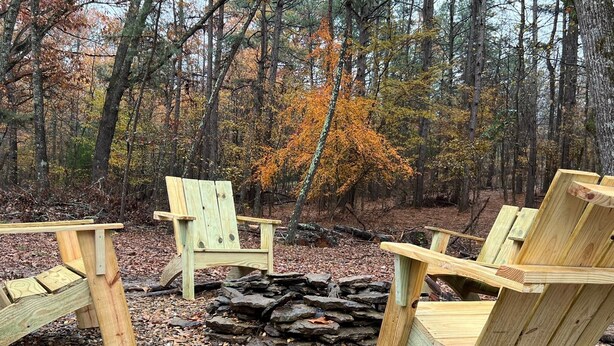 Ein Adirondack-Stuhl aus Holz in einem Wald, mit einem Feuerkorb aus Stein.
