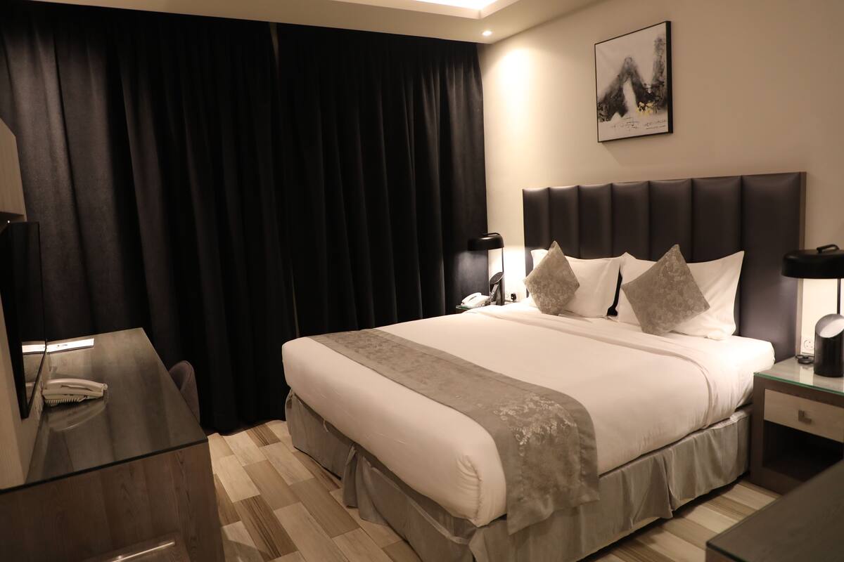 Ein Hotelzimmer mit einem großen Bett, einem Nachttisch, einem Schreibtisch mit Telefon und einem an der Wand hängenden Kunstwerk.