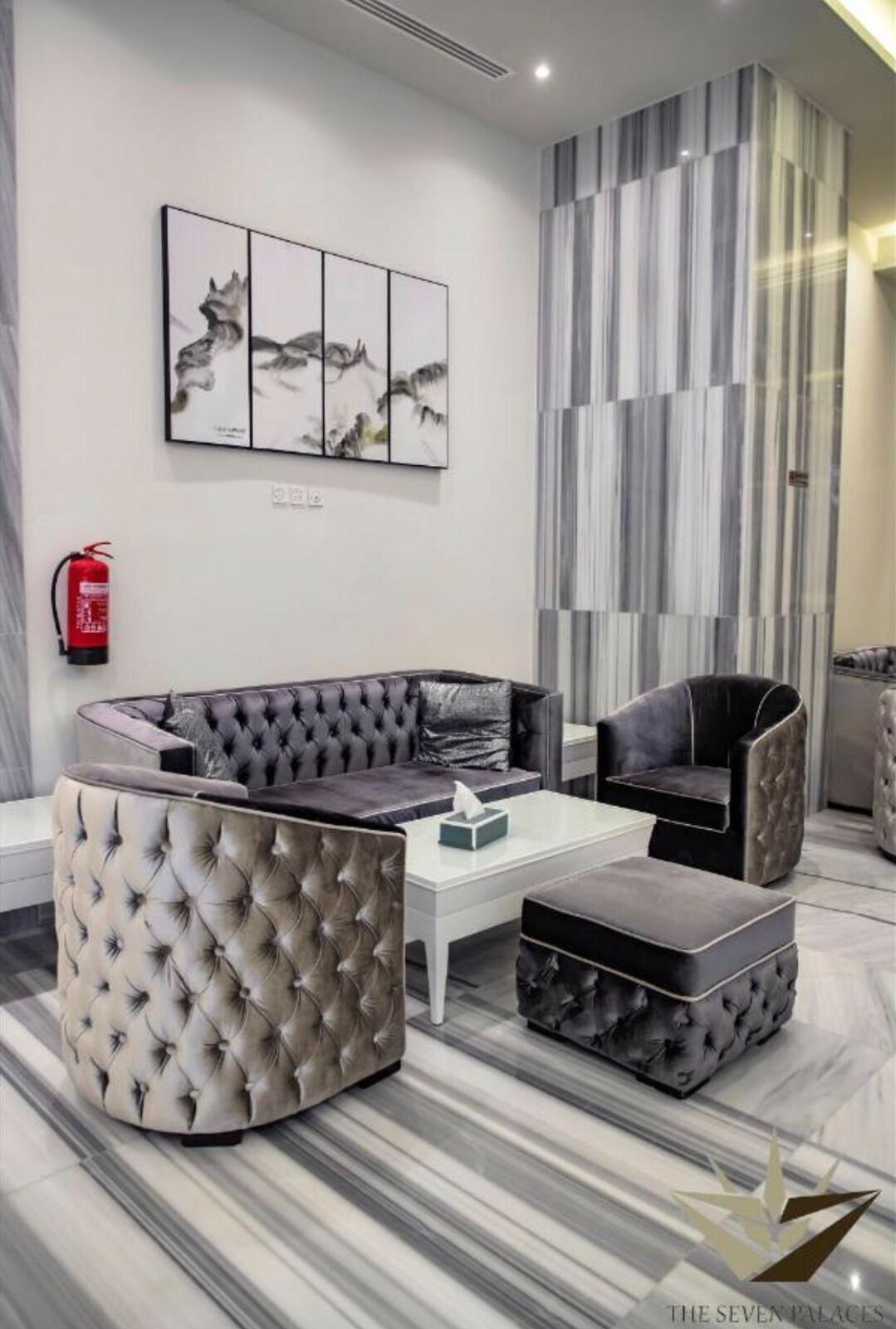A modern living room with a sectional sofa, a chaise lounge, and a coffee table. There is a fire extinguisher mounted on the wall and a framed artwork above the sofa.
