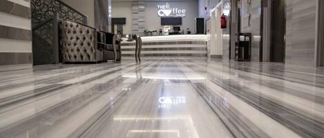 A modern hotel lobby with a marble floor, a reception desk, and a coffee shop named "The Coffee Shop."
