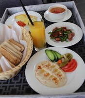 Una bandeja con diversos alimentos para desayuno, incluyendo tostadas, un tazón de sopa, un plato con tomates y pepinos en rodajas, un vaso de jugo de naranja y un plato con salchichas y pimientos.