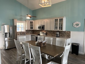 A modern kitchen with white cabinetry, a stainless steel refrigerator, a microwave, and a coffee maker. There is a dining table with white chairs and a ceiling fan above.