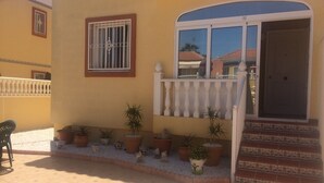 A yellow building with a balcony, potted plants, and a door with steps leading to the entrance.