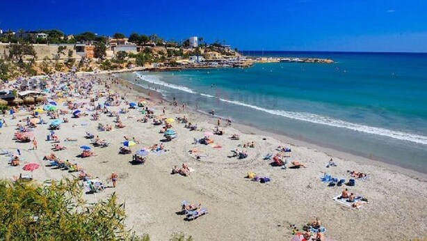 A beach with numerous sunbeds and umbrellas, a clear blue sky, and a rocky coastline.