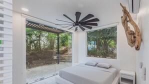 A modern bedroom with a large bed, a ceiling fan, and a view of a garden through a glass door.