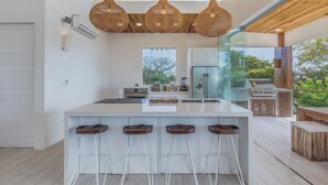 A modern kitchen with a large island, bar stools, hanging wicker light fixtures, and a view of the outdoors.